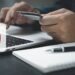 Uma pessoa segura um cartão de crédito enquanto digita em um laptop, simbolizando compras on-line ou internet banking. Um símbolo de pagamento sem contato é sobreposto, refletindo a ascensão dos pagamentos eletrônicos. Um caderno e uma caneta são visíveis em primeiro plano.